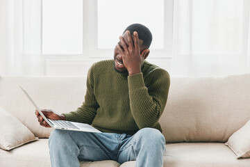 Young Black man expressing frustration while working on a laptop at home, wearing a cozy green...