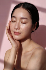 Naklejka premium 頬に手をあて、目を閉じて穏やかな表情を浮かべている黒髪の日本人女性、ピンクの背景、光と影のコントラスト、セルフラブとセルフケア、スキンケアをの重要性を伝える写真、ミニマリスティック雰囲気