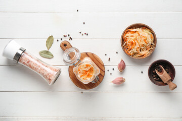 Glass jar, bowl with tasty sauerkraut and condiments on white wooden background. Top view