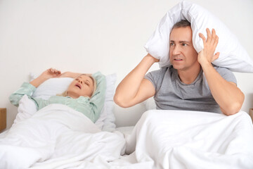 Annoyed mature man with pillow and his snoring wife in bedroom