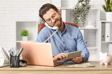 Obraz premium Young businessman with calculator talking by telephone at table in office
