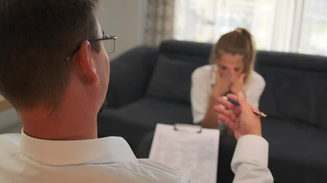 Over shoulder view of the young woman sitting on the couch in the psychotherapist's office, covering her face with her hands, releasing her fears and emotions, and expressing deep emotional tension.
