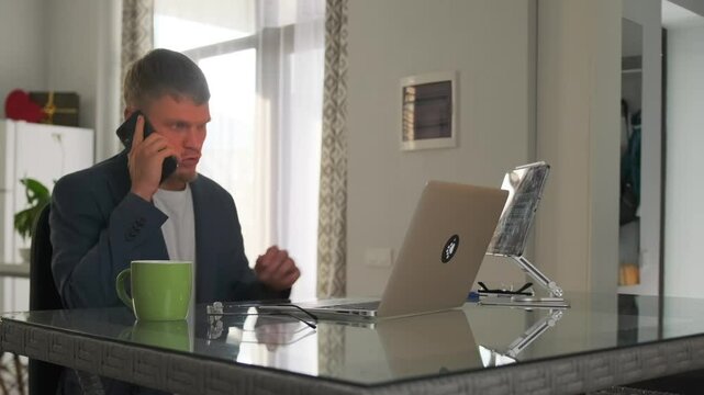 The young businessman in a suit, working from home, sits at his laptop during a work call, discussing with colleagues and clarifying details of the budget and operations.