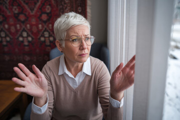Senior woman stares out suspiciously through her blinds, her fingers separating them