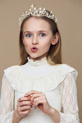 Young girl with a crown in a vintage lace dress, expressing surprise against a soft beige...
