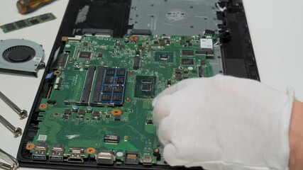 A technician wearing white gloves is carefully handling the motherboard of a laptop during a repair process, ensuring precision and cleanliness in a workshop.
