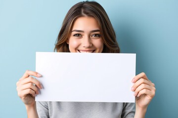 Naklejka premium A cheerful woman holding a blank paper sign, set against a light blue background, smiling brightly at the camera.