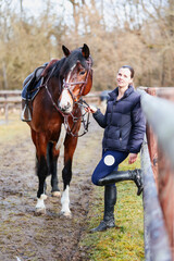 Obraz premium A young woman stands with her saddled horse model between the wooden fences of the outdoor paddocks in winter nature.