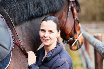 A young woman stands with her saddled horse model between the wooden fences of the outdoor paddocks...