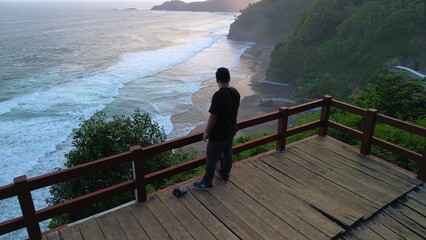  A man standing in the top of hill in coastline with hills, cliffs, trees, beach sand and waves from the sea and ocean at Lampon Beach, Kebumen, Central Java, Indonesia