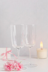 Two empty transparent wine glasses or champagne flute served on white wooden table with pink carnation flowers glowing candle light and surprise gift box for celebration of engagement proposal holiday