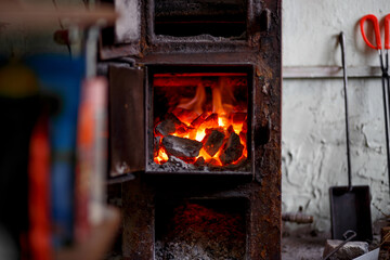 A rustic stove with a burning fire, a coal and firewood shovel and tongs standing nearby. Heating in a private house, fireplace, fire safety.