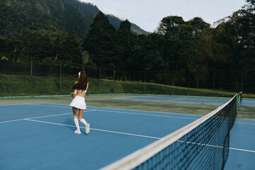 Fototapeta premium Athletic young woman in white tennis outfit practicing on a blue court surrounded by lush green trees, showcasing focus and determination in a serene setting