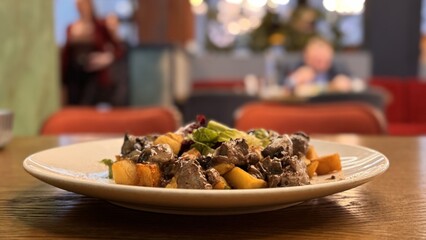 Warm salad with chicken liver in cream sauce with fried potatoes, mushrooms and mixed green salad in a white porcelain plate on a brown wooden table in an elite restaurant. Close-up