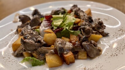 Warm salad with chicken liver in cream sauce with fried potatoes, mushrooms and mixed green salad in a white porcelain plate on a brown wooden table in an elite restaurant. Close-up