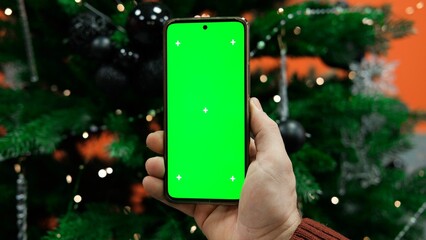 Young man holding smartphone with green screen at work in office against christmas tree background. Vertical mockup of mobile phone. Man using phone with blank green screen. Smartphone with chromakey