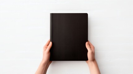 Hands holding a blank black notebook on a white surface during daylight