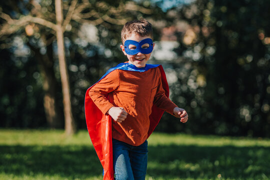 Young boy in superhero costume runs joyfully through green grass in sunny park during afternoon playtime