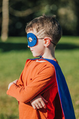 Naklejka premium Young child in superhero costume poses confidently in a sunny park with a green background