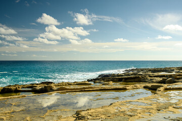 A stunning view of coastal cliffs with layers of golden sandstone overlooking a vibrant turquoise sea. The sunlight highlights the rugged textures of the rocks.