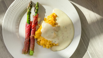 A gourmet breakfast dish featuring roasted asparagus wrapped in prosciutto with creamy scrambled eggs and hollandaise sauce, perfect for Easter brunch concepts