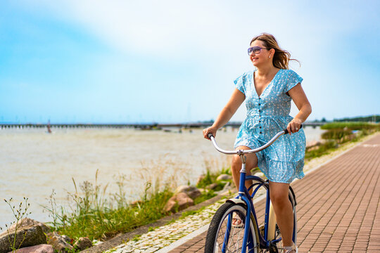 Active summer holidays. Mature woman wearing dress riding blue cruiser bike on bicycle path on seafront on sunny day. Front view