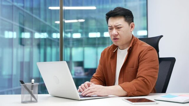 Shocked stressed asian businessman reads bad news on laptop while sitting at desk at workplace in business office. Frustrated worried worker is upset while reviewing negative information on a computer