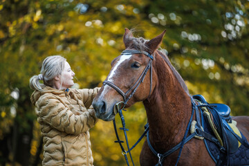 Woman preparing her bay horse, animal harness and saddle for recreational horseback riding in nature
