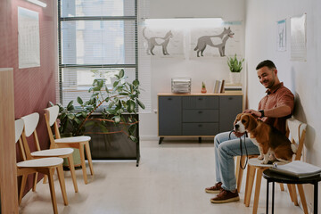 Man sitting in veterinarian's waiting room with dog on lap, surrounded by chairs and decor. Various...
