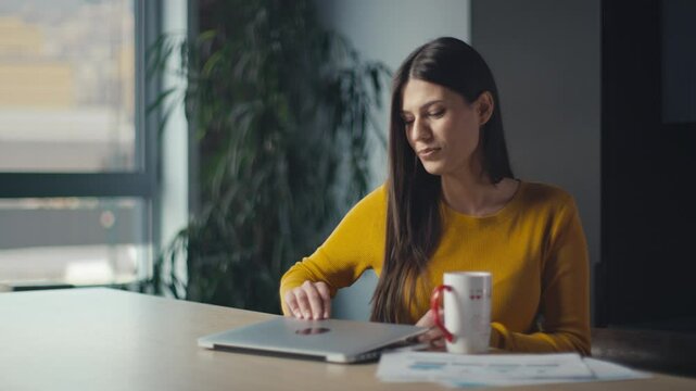 Woman in a yellow sweater waves goodbye during an online call, holding a mug, then closes her laptop and stands up. Concept of remote work, virtual meetings, or ending online communication