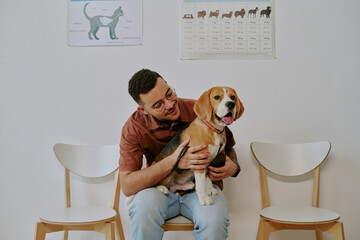 Young man smiling while holding a beagle in his lap, sitting on a chair in front of white wall with...