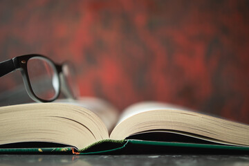 Open book lying on tabletop and glasses lying on book, red blurry wall in background. Space for text, design.