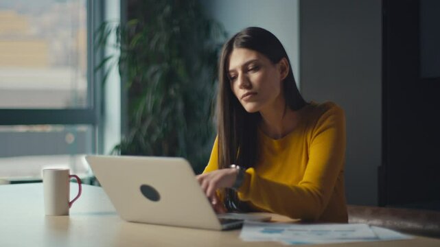 Young woman smiling while working on a laptop in a modern office, wearing a yellow sweater, radiating happiness and focus. Concept of positivity, productivity, and professional lifestyle