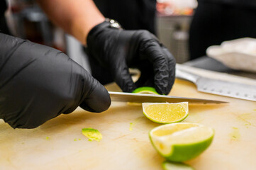Close-up of a chef's hands with black gloves meticulously slicing a fresh lime on a white cutting...