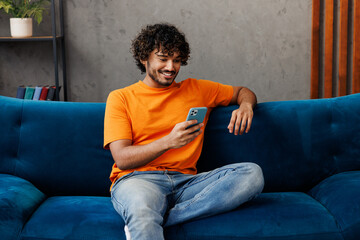 Young Indian man wear orange casual clothes sits on blue sofa couch hold in hand use mobile cell...