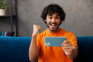 Young winner man wear casual clothes sits on blue sofa couch use play app on mobile cell phone hold gadget for pc video games stay at home hotel flat rest relax spend free time in living room indoor.