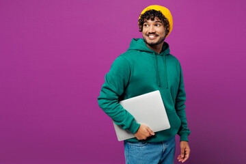 Young happy Indian IT man he wear green hoody yellow hat casual clothes hold closed laptop pc computer look aside on area mock up isolated on plain purple background studio portrait Lifestyle concept