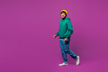 Full body side profile view young smiling happy fun Indian man he wear green hoody yellow hat casual clothes walk go look camera isolated on plain purple background studio portrait. Lifestyle concept.