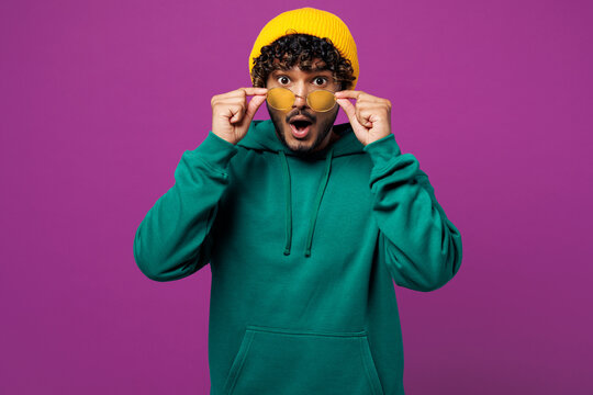 Young happy shocked surprised impressed Indian man he wearing green hoody yellow hat sunglasses casual clothes lower glasses isolated on plain purple wall background studio portrait Lifestyle concept