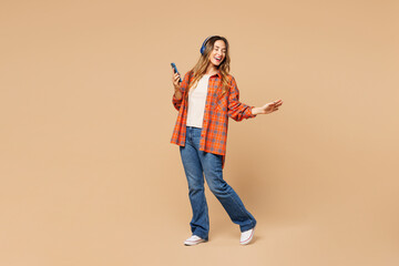 Fototapeta premium Full body young fun woman she wear orange shirt casual clothes listen to music in headphones use mobile cell phone isolated on plain pastel light beige background studio portrait. Lifestyle concept.