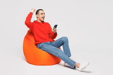 Full body young middle eastern man he wear red hoody casual clothes sit in bag chair hold in hand use mobile cell phone do winner gesture isolated on plain solid white background. Lifestyle concept.