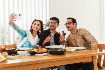 An Indian family three people, father, daughter and son are taking a selfie at a dining table, surrounded by traditional food, smiling and enjoying a joyful moment together. Special meal for family.