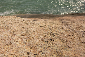 Close-up of a textured rock surface near the water. The rock looks uneven and weathered, with barely noticeable color transitions. There is a shimmering ocean in the background.