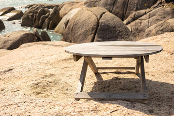 There is a wooden table on the stone surface overlooking the ocean. In the background, waves crashing against the shore and large boulders. Beach theme and recreation.