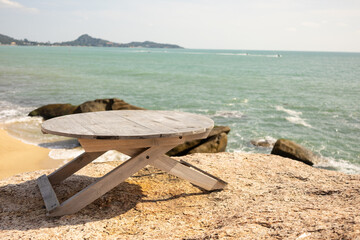 There is a wooden table on the stone surface overlooking the ocean. In the background, waves crashing against the shore and large boulders. Beach theme and recreation.