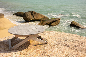 There is a wooden table on the stone surface overlooking the ocean. In the background, waves crashing against the shore and large boulders. Beach theme and recreation.