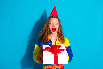 Colorfully dressed young woman holding a gift box with a red ribbon, wearing a party hat,...