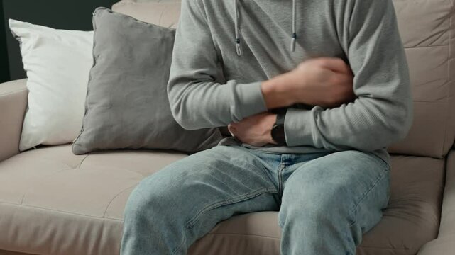 Close-up of man sitting on a couch at home, clutching his stomach, suffering from abdominal pain or cramps. Person suffering from health issues, digestion problems or food poisoning