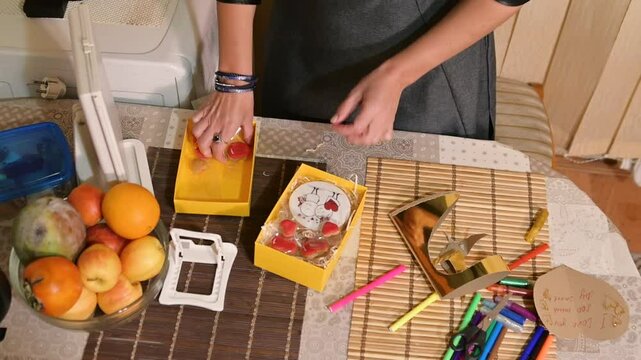 A woman is wrapping a present in a yellow gift box. The box contains heart-shaped cookies.
