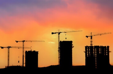 Naklejka premium Silhouettes of tower cranes constructing a new residential building at a construction site against sunset background.